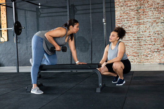 Latina client lifting dumbbell on bench with African American woman coach crouching, copy space