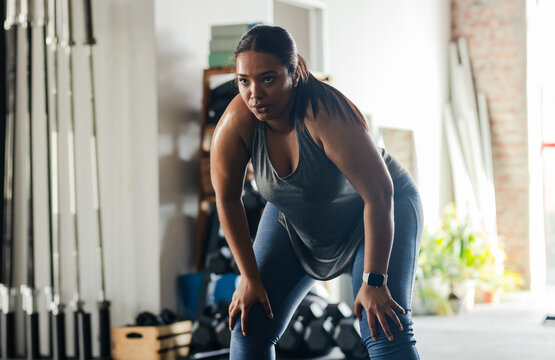 African American woman panting in workout attire, leaning on knees at gym near weight bars