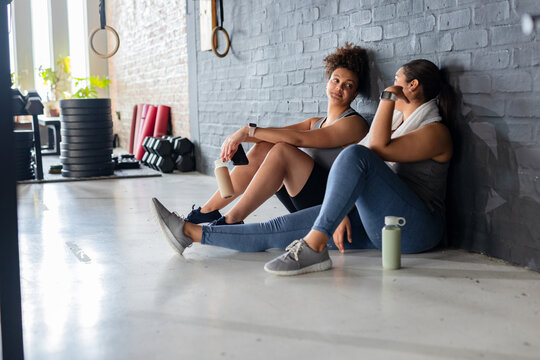 Diverse female friends resting at fitness studio holding smartphone and water bottles, copy space - Powered by Adobe