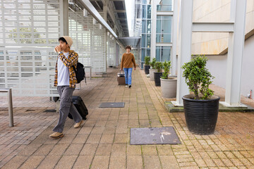 Diverse male friends walking by glass building, pulling suitcases and sipping coffee, copy space