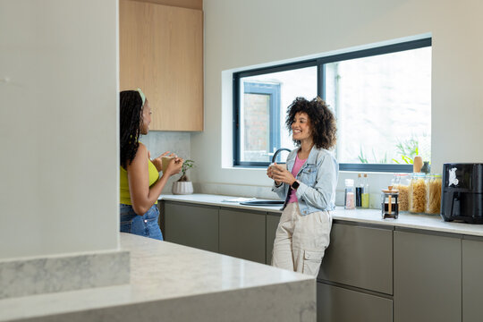 Diverse female friends chatting, holding mugs near island with air fryer in kitchen, copy space