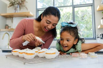 Hispanic mother and daughter piping white icing onto orange cupcakes in home kitchen with sprinkles