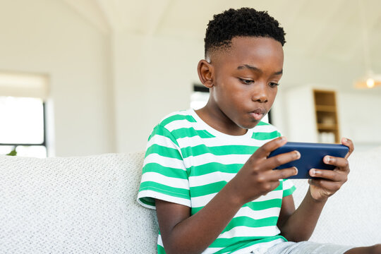 African American boy holding smartphone, focusing on screen, sitting on sofa at home, copy space