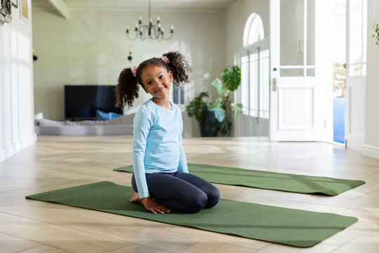 African American girl kneeling on green yoga mat in living room beside arched doors and plants
