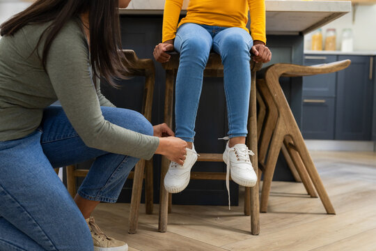 African American mother and daughter kneeling tying shoelace on wooden stool in residential kitchen