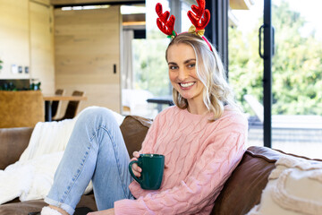 Woman sitting sideways on sofa in living room wearing sweater antler headband holding green mug