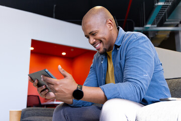 African American man using tablet on gray sofa in office lounge checking smartwatch by coffee cup