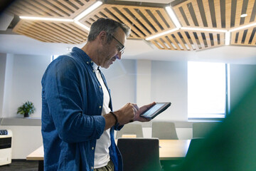 Mature adult man holding tablet and reviewing at wooden table in conference room with grid ceiling