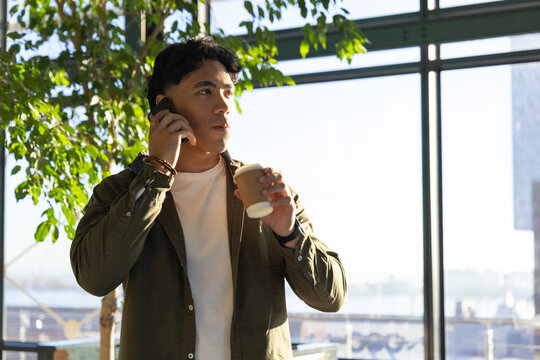 Asian man talking on smartphone while holding disposable coffee cup near leafy plant in cafe lobby