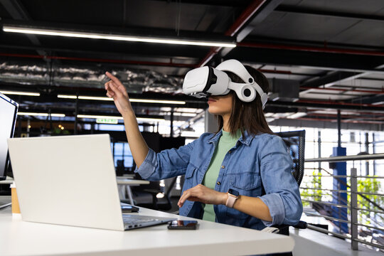 Laptop and smartphone resting on white office desk with linear LED lighting, VR headset, coffee cup