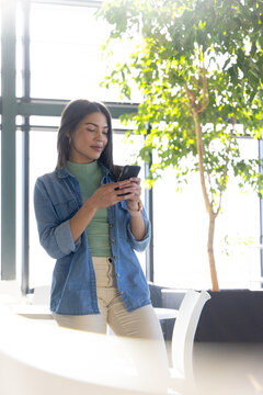 Asian Indian woman leaning against white table in office lounge using smartphone, copy space