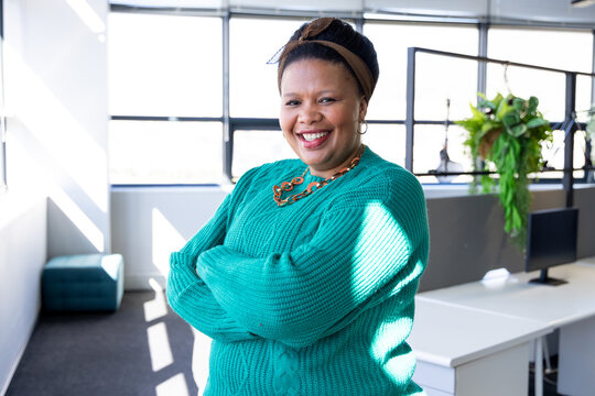 African American woman standing wearing teal sweater in office near grid windows with desk, ottoman