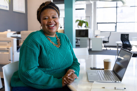 African American woman sitting at modern office desk smiling and working on laptop with coffee cup