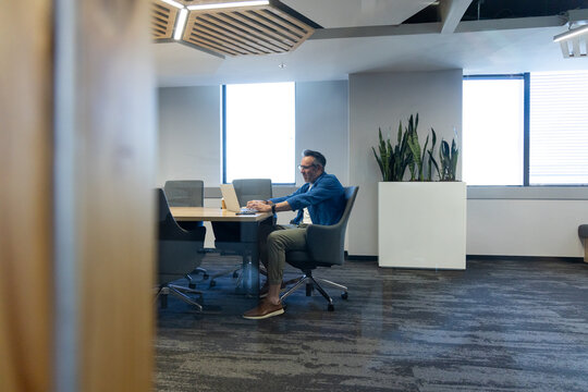 Laptop is resting on conference table in office with lights and tall green plants, copy space