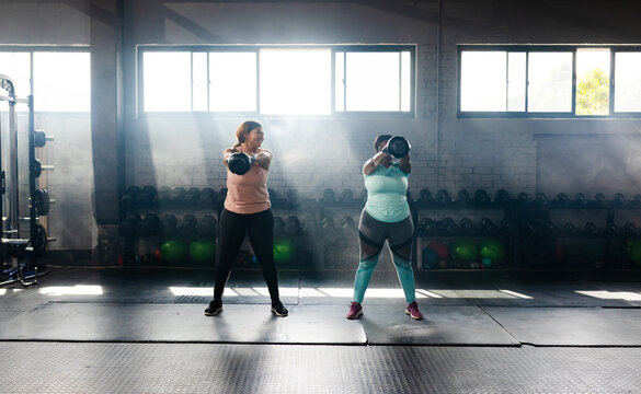 Diverse female workout partners holding kettlebells and training in gym with dumbbell rack and mats