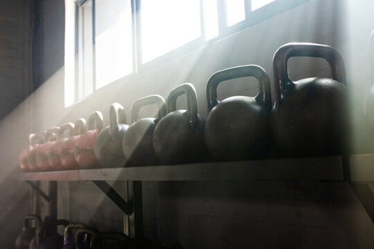 Cast-iron kettlebells sitting on metal shelf under high windows, sunlight beams highlighting dust