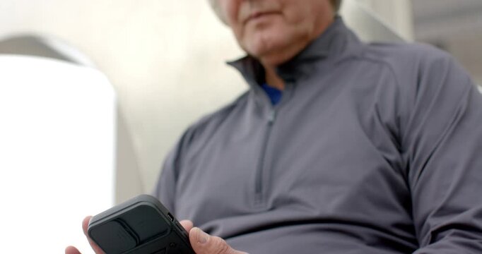 Senior man tapping and swiping rugged smartphone while unzipping jacket and leaning in lounge area