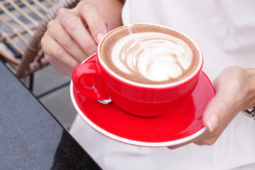 Female tourist hand holding a red cup and saucer, hot coffee latte art at a coffee shop while relaxing in the morning.

