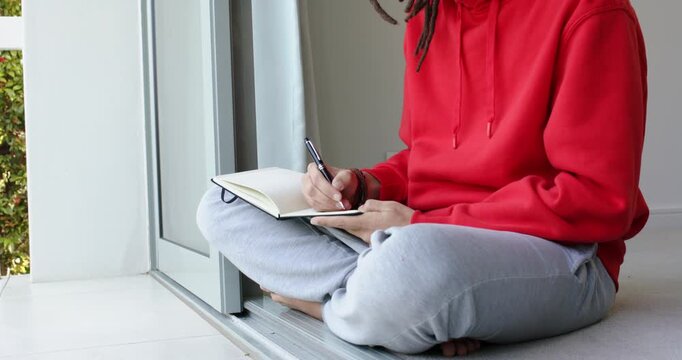 Writer in hoodie is pressing pen tip and writing crosslegged at sliding door threshold with curtain
