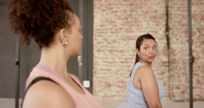 Diverse female friends holding squat turning head to acknowledge partner's glance at gym with rack