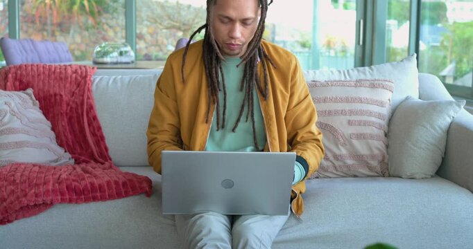 Man sitting on sofa opening laptop, typing, smiling at funny content, pausing on noise, refocusing