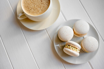 White macarons filled with cream and a cup of frothy coffee resting on a light wooden table, creating a serene and inviting scene for a sweet snack or breakfast moment