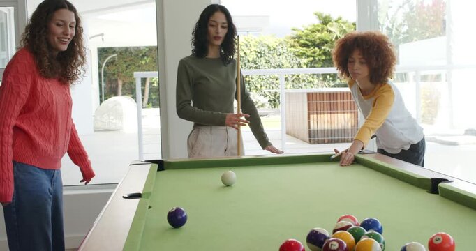 Three female friends lining shot, striking ball, cheering around pool table in home recreation room