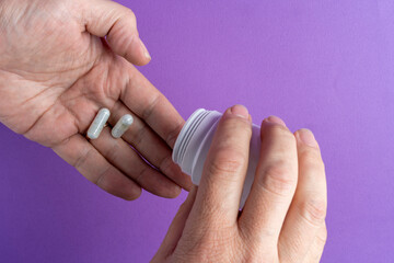 Person pouring white capsules from bottle into hand on purple background, close-up healthcare concept