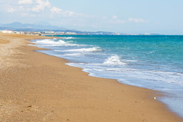 A serene empty beach with turquoise water and white foam on wet sand, mountains and hotels visible in the distance. A peaceful pre-season vista. Sorgun, Antalya, Turkey.