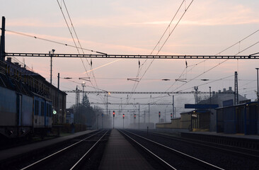 Obraz premium Atmospheric view of a railway station at dawn with pink and blue sky, illuminated red signals, overhead electric lines, and a parked train on the tracks