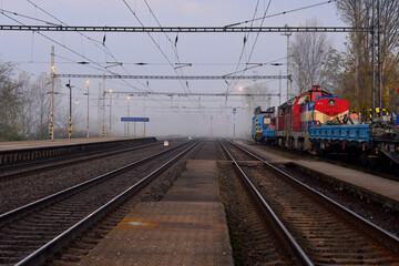 View down railway tracks at a foggy station with a maintenance train parked on the side and complex overhead electric lines