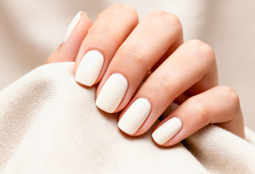 A close-up image depicts a hand with short, neatly shaped nails, featuring a simple and elegant manicure on a light neutral background, capturing subtle skin texture and textile detail