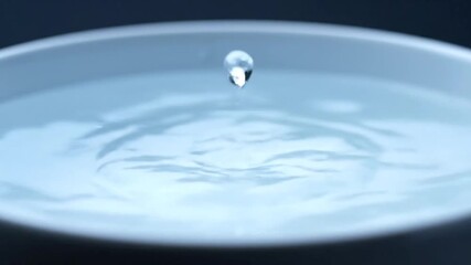 Close-up of a single water droplet falling into clear blue water creating ripples and splashes within a white bowl under soft studio lighting