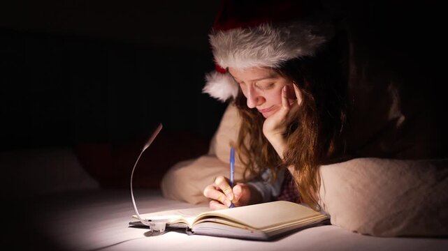 Young woman in a santa hat lies in bed at night, writing in a notebook by a soft lamp penning a christmas letter, wish list, or private holiday diary in cozy surroundings