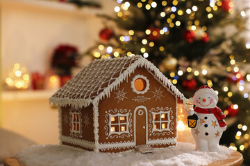 Naklejka premium Beautiful gingerbread house, snowman figure and artificial snow on table against blurred lights indoors, closeup. Christmas atmosphere