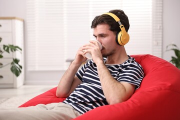 Young man in headphones with cup listening music on bean bag chair at home