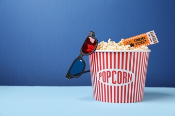 Tasty popcorn in paper cup, movie ticket and 3D glasses on table against blue background, space for text