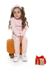 Cute little girl in headband sitting on pouf and gift box against white background