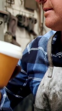Vertical video: Reaching for coffee cup, apron-clad metalworker drinking to take break at workshop