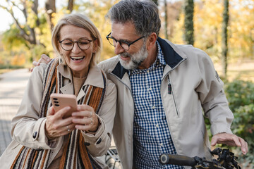 Happy senior couple looking at a smartphone together in a park, smiling and enjoying a cheerful moment on an autumn day.