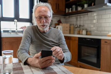 Elderly man sitting at a kitchen table, focused on his smartphone, with natural light illuminating...