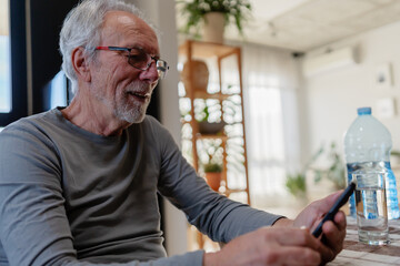 Smiling senior man sitting at a kitchen table, using his smartphone with a glass of water beside him in a cozy home setting