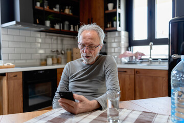Smiling senior man sitting at a kitchen table, using his smartphone with a glass of water beside him in a cozy home setting