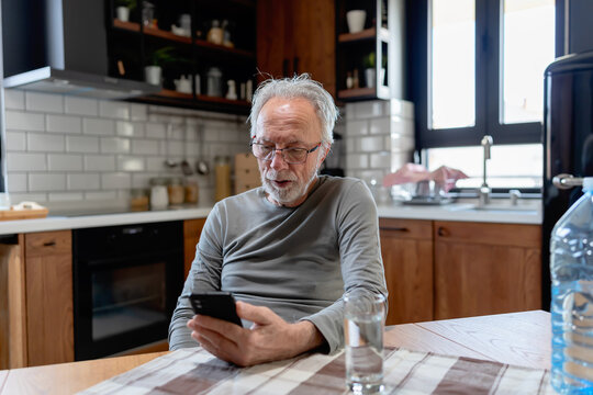 Smiling senior man sitting at a kitchen table, using his smartphone with a glass of water beside him in a cozy home setting