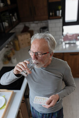 Elderly man in his kitchen taking daily medication, holding a pill and drinking water as part of his morning health routine