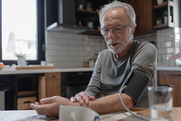 Senior man sitting at a kitchen table using a digital blood pressure monitor, checking his health...