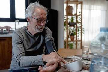 Senior man sitting at a kitchen table using a digital blood pressure monitor, checking his health...