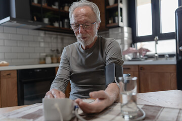 Senior man sitting at a kitchen table using a digital blood pressure monitor, checking his health in a bright, cozy home environment