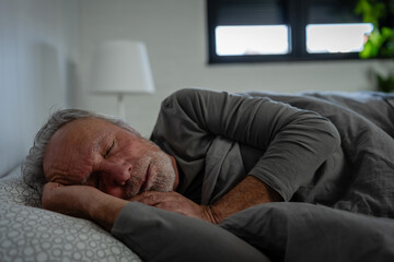 Elderly man sleeping peacefully in bed, resting on his side under a gray blanket in a softly lit bedroom