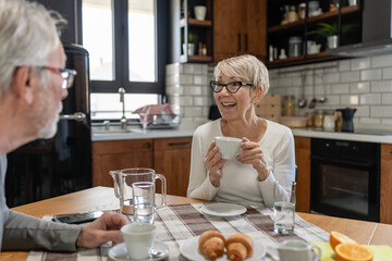 Smiling senior woman enjoying coffee at the breakfast table with her partner in a cozy kitchen setting